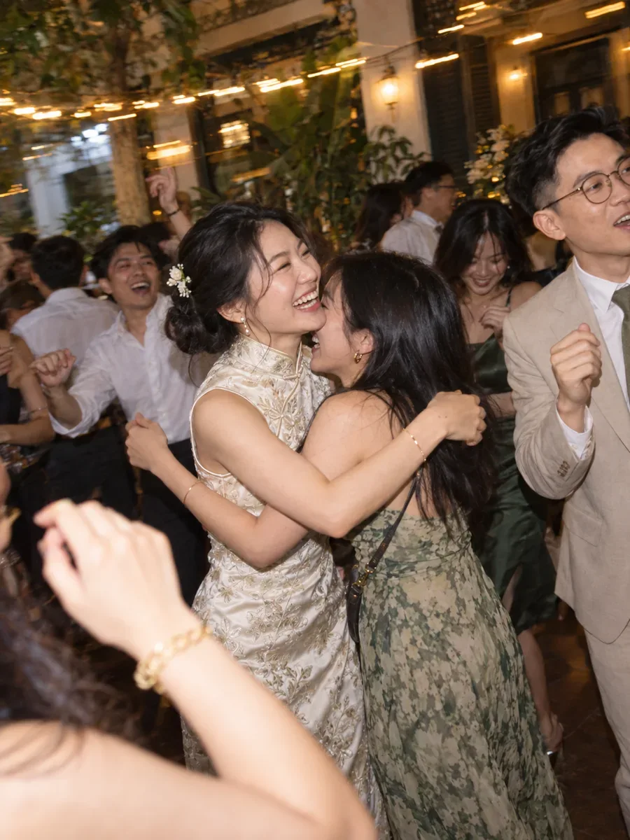 Illustrative wedding scene of a bride embracing a friend among guests under string lights.